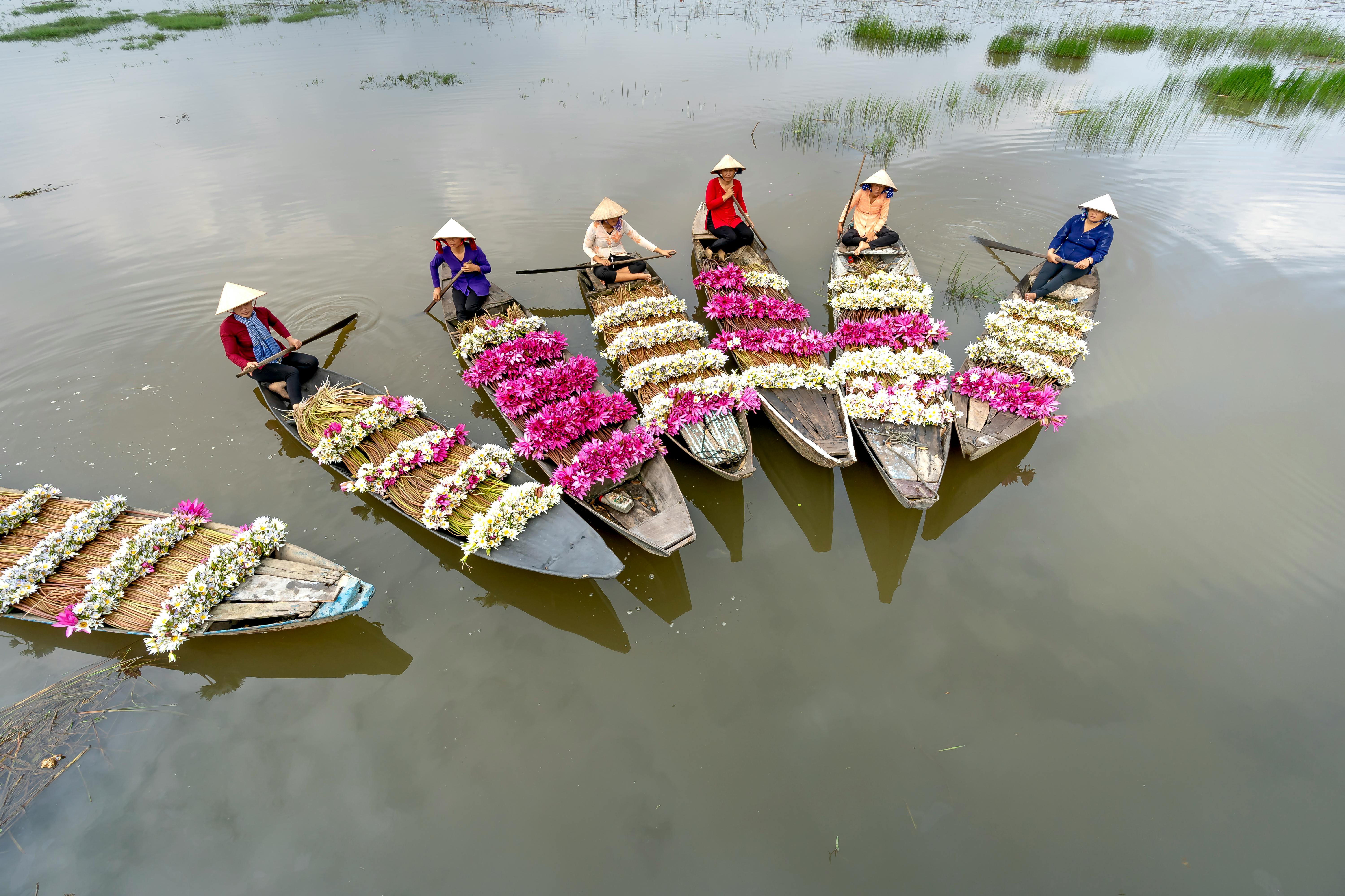 Aerial view of boats with vibrant flowers and women in conical hats on a river in Vietnam.