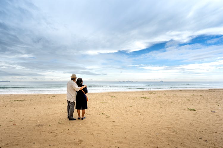 Back View Of A Senior Couple Standing On A Sandy Beach And Looking At Sea