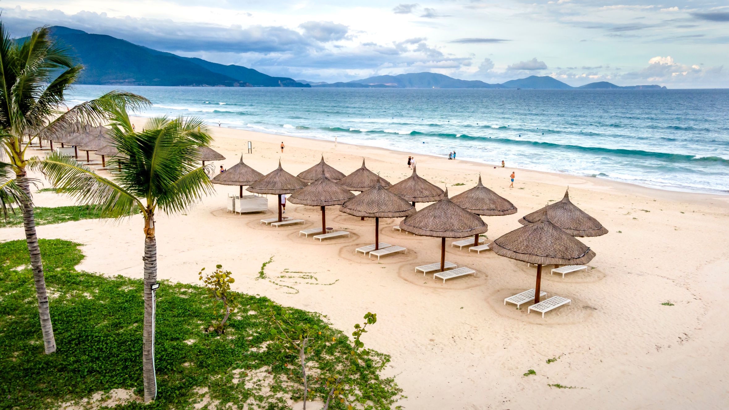 Serene sandy beach with palm trees and umbrella huts under a cloudy sky.