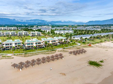 Aerial shot of beachfront with umbrella huts, luxury resort, and mountains in the background.