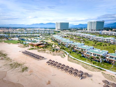 A stunning aerial view of a modern beachfront resort with umbrella huts and lush greenery.