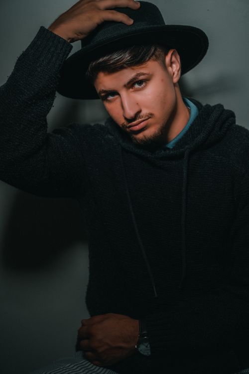 Free A fashionable man in a black sweater and fedora hat posing confidently against a dark background. Stock Photo
