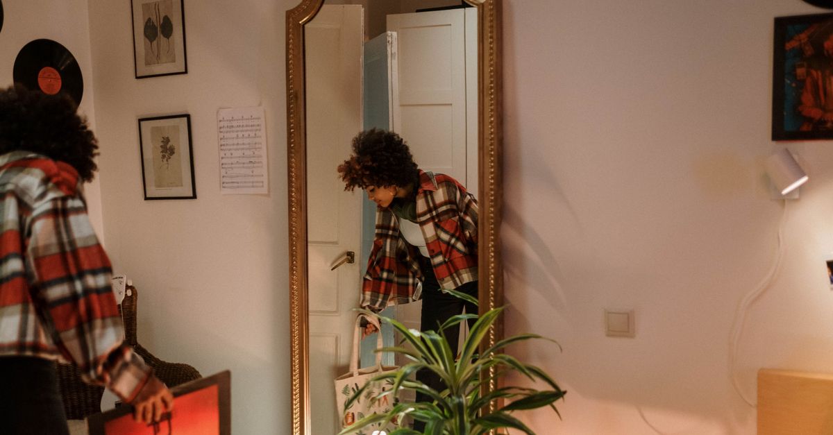 Uma mulher com cabelo afro em traje xadrez segurando uma bolsa, refletindo em um espelho vintage em casa.