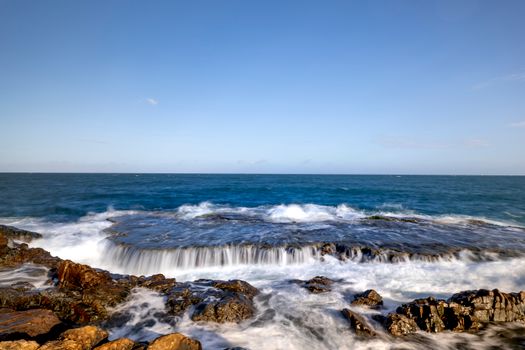 A vibrant coastal scene featuring powerful waves crashing onto rocky shores under a clear blue sky.