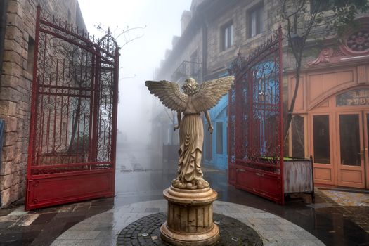 A serene misty street featuring an elegant angel statue flanked by ornate gates.