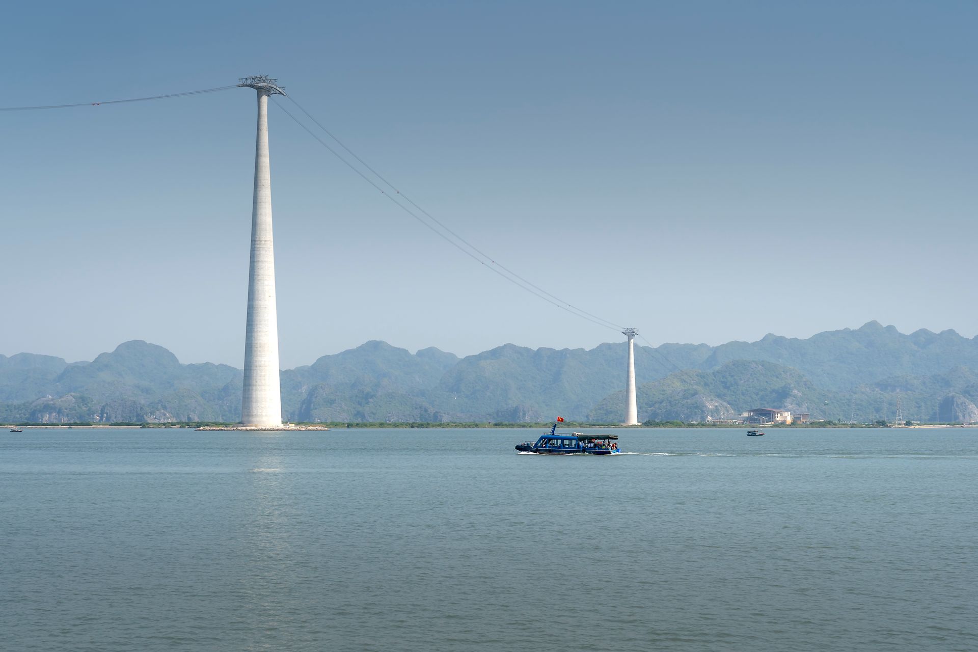Scenic view of cable car tower over Halong Bay with a boat in foreground and hills in backdrop.