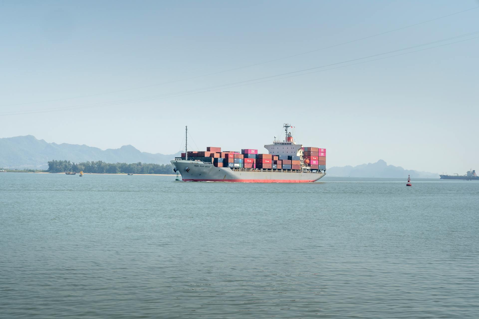 A large container ship sails in calm waters under a clear sky, mountains in background.