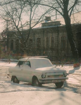 A classic car covered in snow parked by an old building during winter.