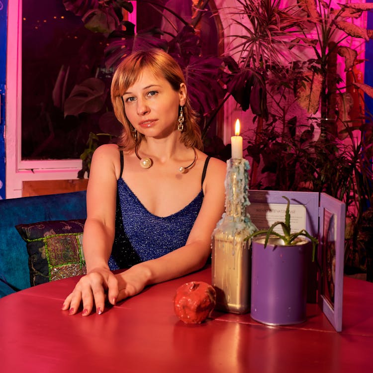 Gorgeous Young Woman Sitting At Table In Dark Restaurant And Looking At Camera