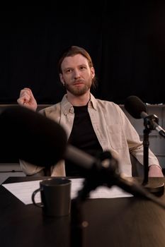 Bearded caucasian man in beige shirt podcasting in a studio with microphones.