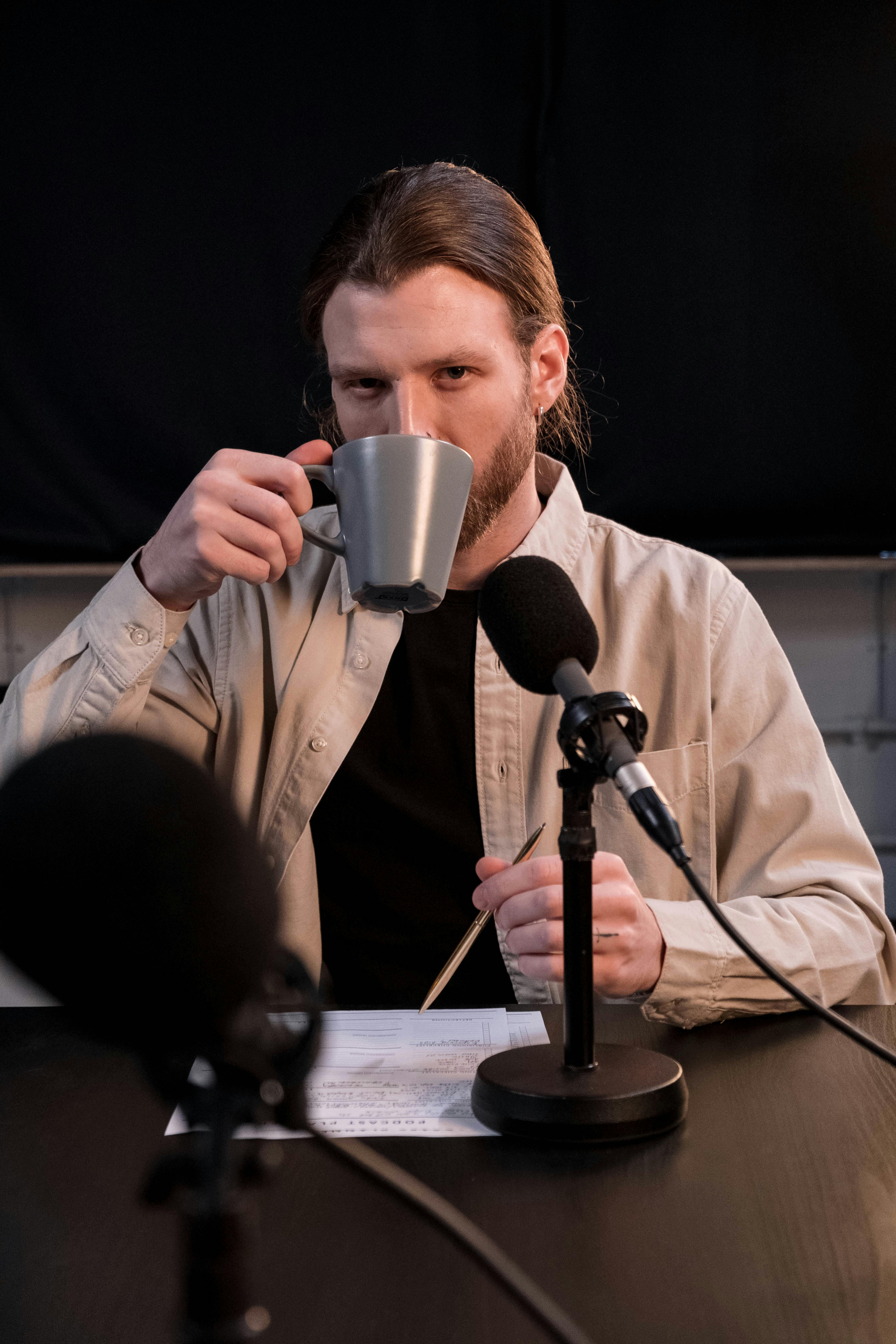 Adult man sipping coffee while podcasting with two microphones indoors.