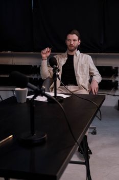 Man in a studio sitting at a table for a podcast interview with microphones.