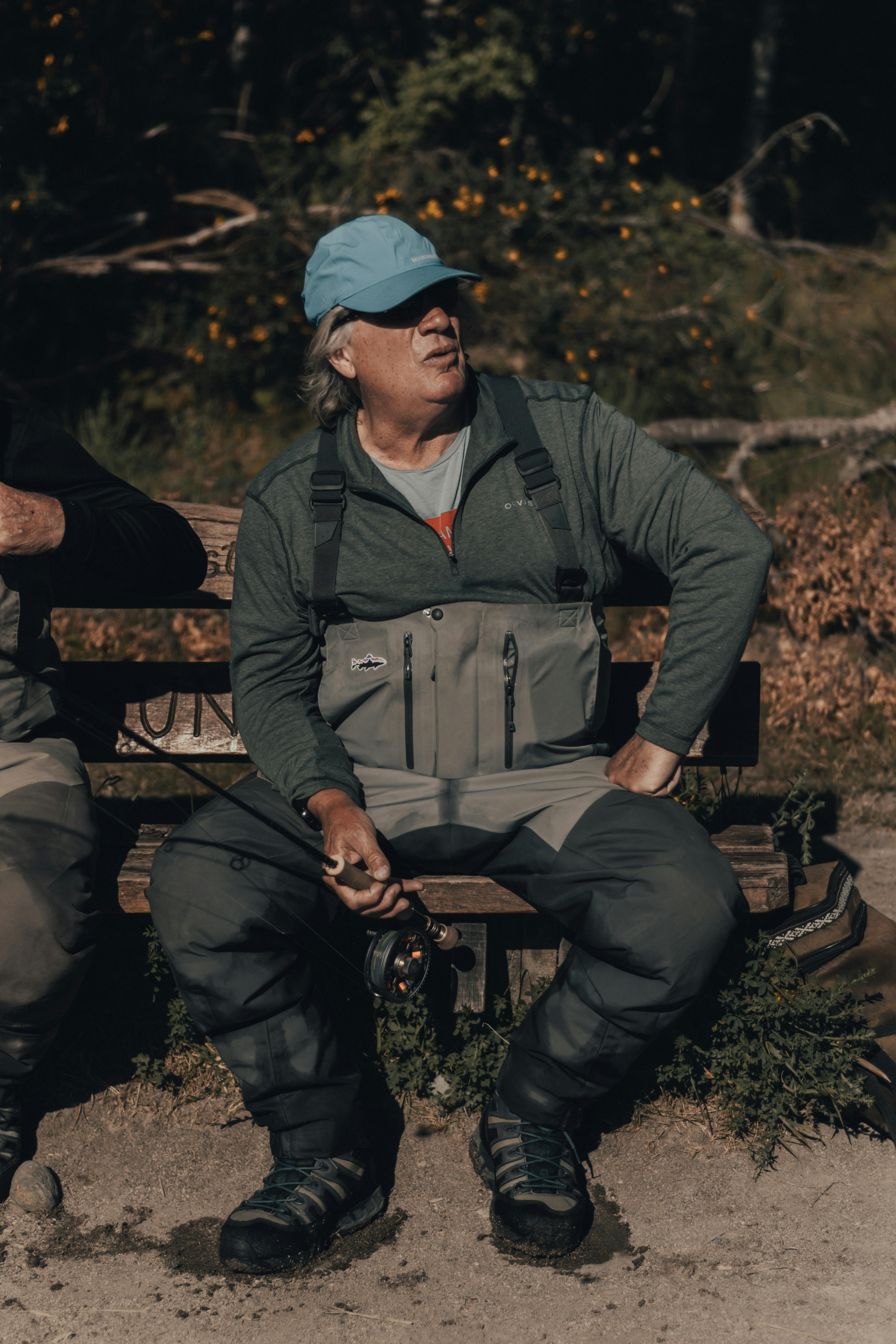 Elderly man in fishing gear sitting on a bench outdoors, holding a rod and looking relaxed.