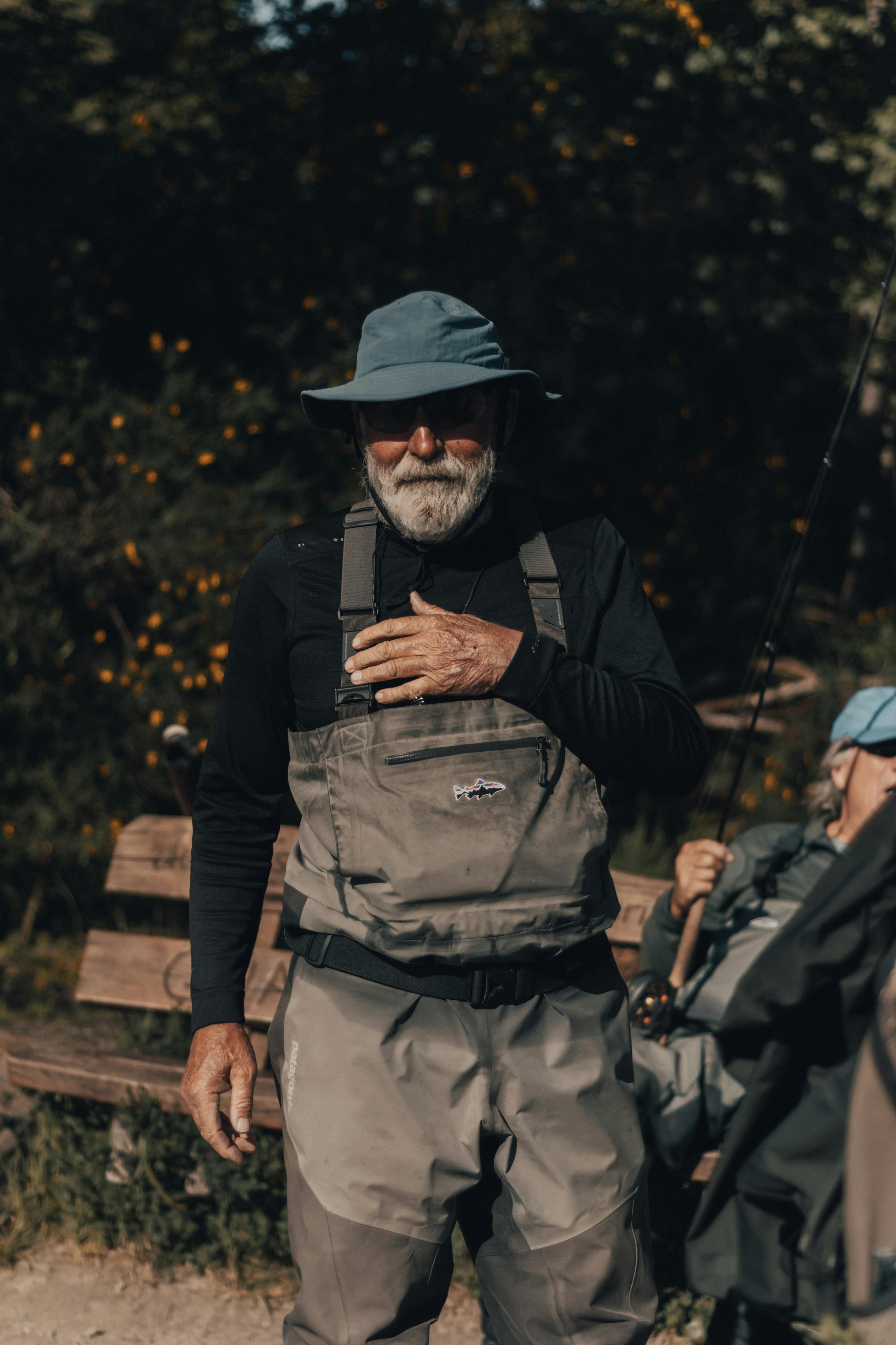 Senior man wearing a bucket hat and overalls, standing with a fishing rod outdoors.