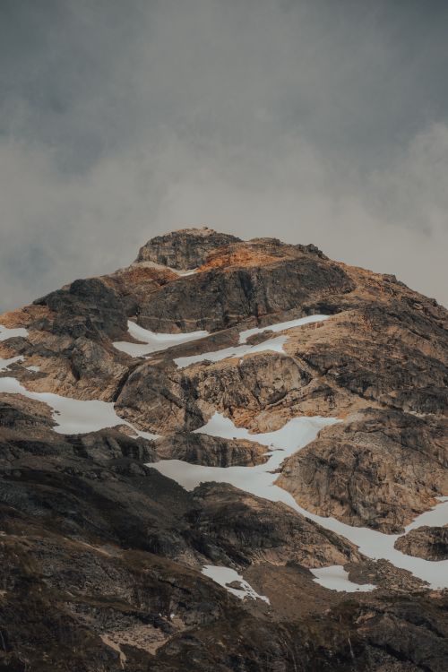 Free Vertical shot of a rugged, rocky mountain peak with snow patches under a cloudy sky. Stock Photo