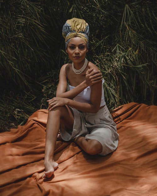 Full body of fashionable African American female wearing authentic headdress looking at camera while sitting on blanket near green plants in nature