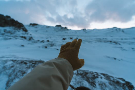 A gloved hand reaches out toward a snowy mountain landscape under a cloudy sky.