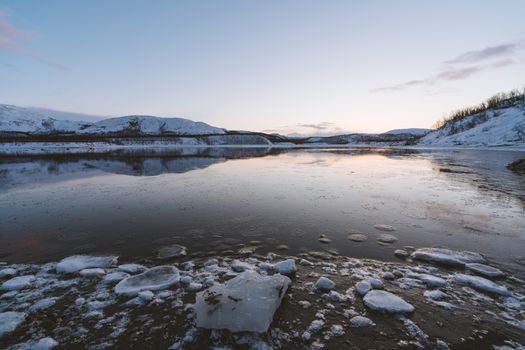 A tranquil frozen lake surrounded by snow-covered hills under a clear winter sky at sunset.