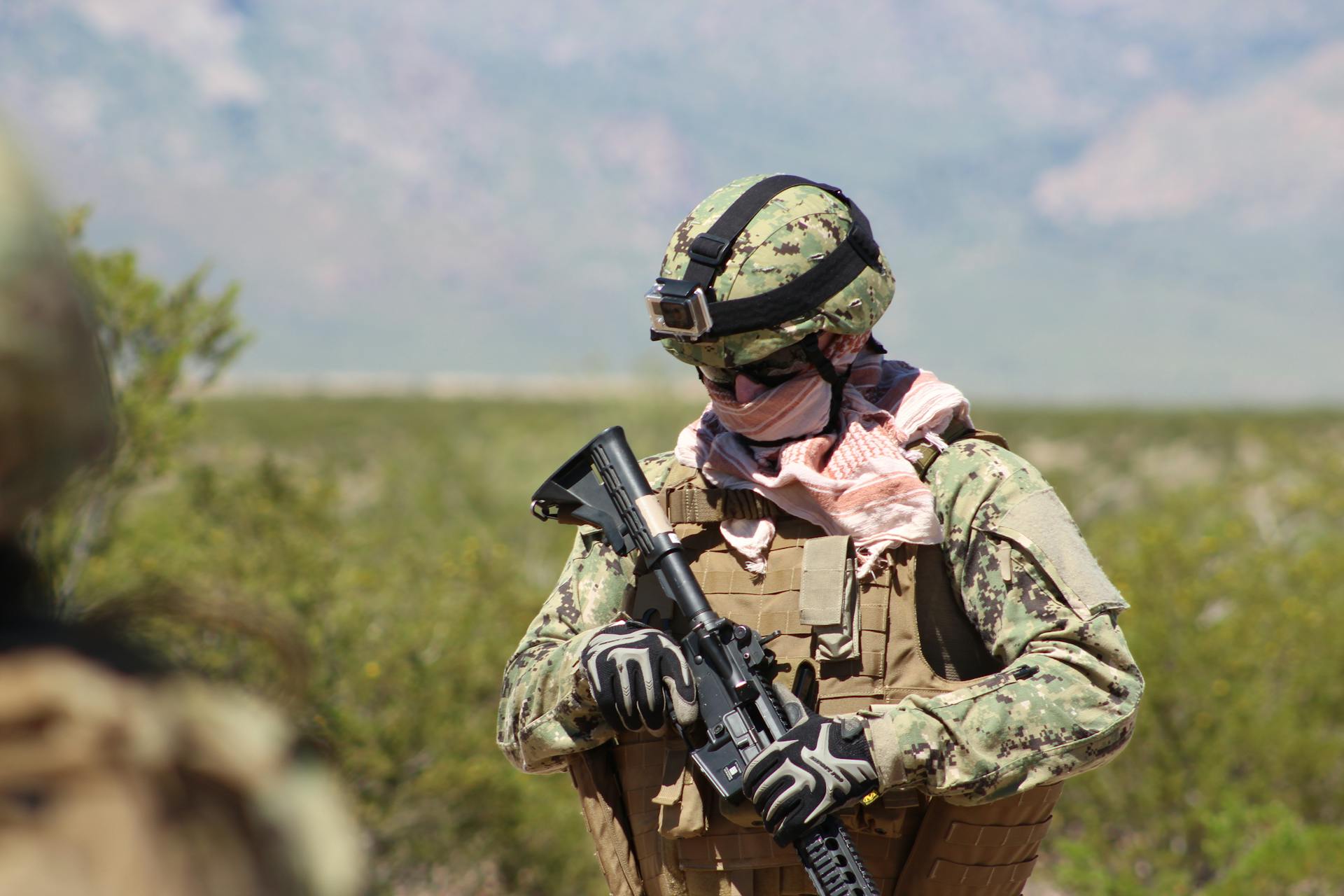 Armed soldier in camouflage gear holding a rifle, positioned in an outdoor setting.