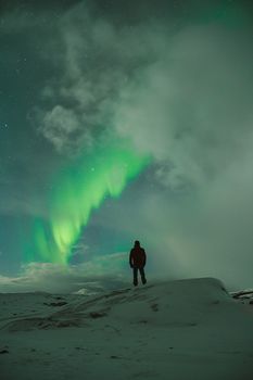 A solitary figure stands under the vibrant aurora borealis against a starry winter sky.