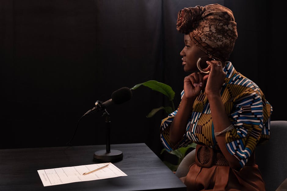 African American woman in studio podcasting, wearing a stylish headwrap and colorful attire, adjusting earring.