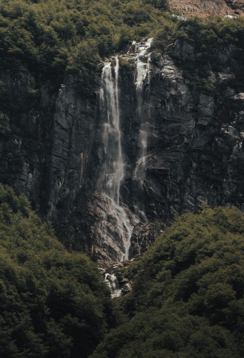 Free A stunning waterfall cascades down a rocky mountain surrounded by lush greenery, creating a serene natural scene. Stock Photo