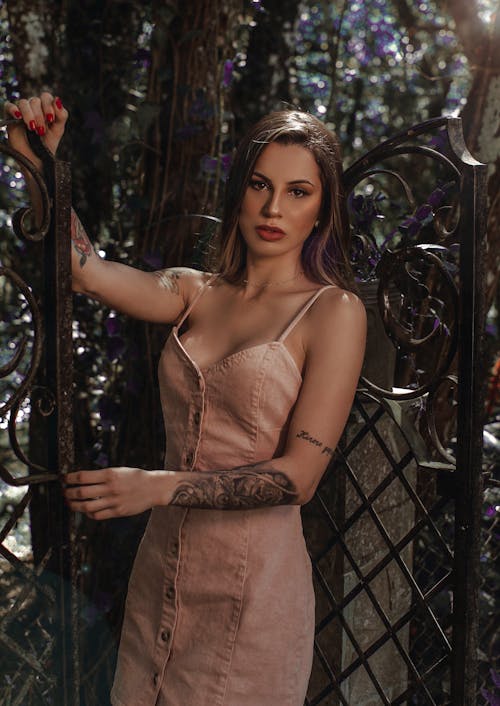 Free Elegant female with red lips and long hair looking at camera while leaning on metal gate of yard Stock Photo