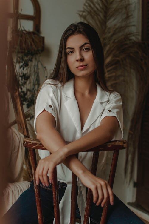 Young female with dark long hair in stylish clothes looking at camera on timber chair