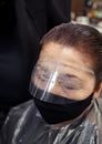 From above of middle aged female with eyes closed in protective mask dying hair in salon
