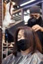 Professional anonymous male in hat dying short hair of elderly female in black protective mask