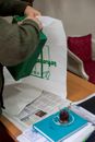 Crop anonymous male putting green box in package near wooden table with glass with drink on saucer and notebook with documents near newspaper in workplace