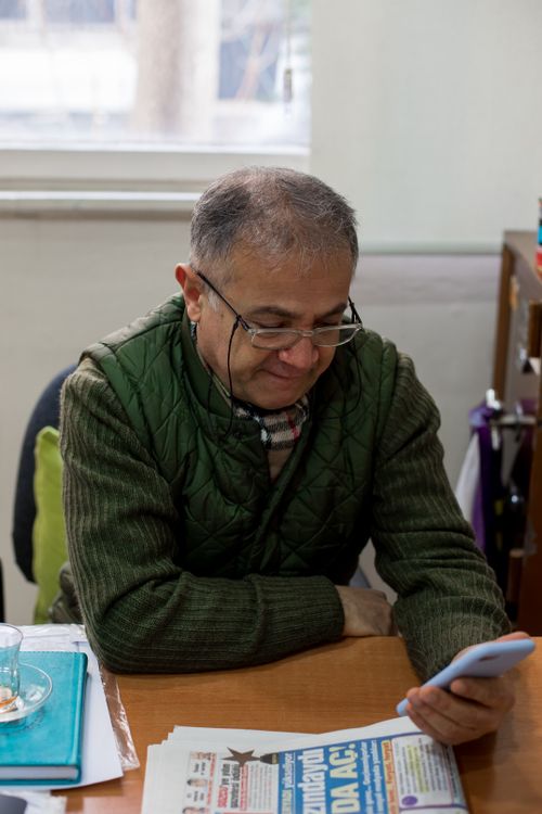 Free Serious middle aged male in eyeglasses and casual clothes sitting at table while surfing on cellphone near newspaper and book next to window in light workspace Stock Photo