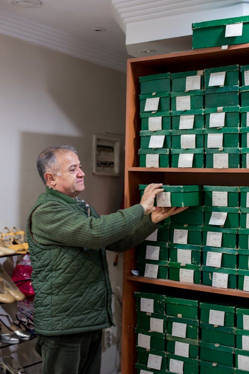 Positive middle aged man in casual outfit putting boxes with shoes on shelves while standing in bright shoe shop near and looking away