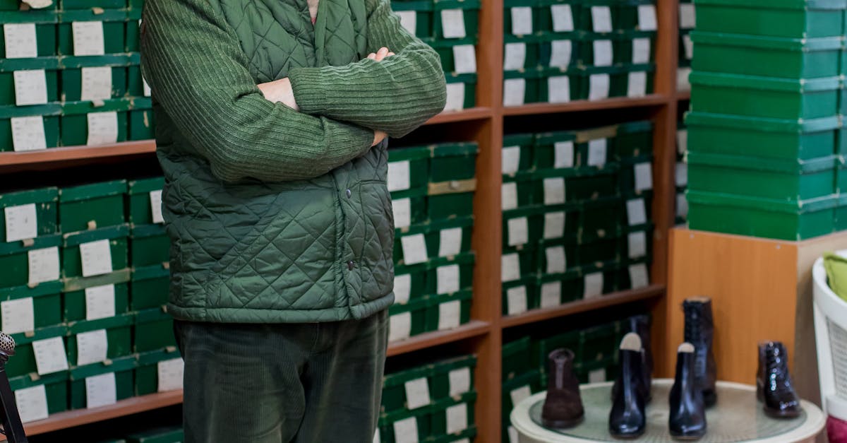 Full body of calm ethnic male customer in uniform standing with crossed arms in shoe store against shelves with boxes and looking at camera