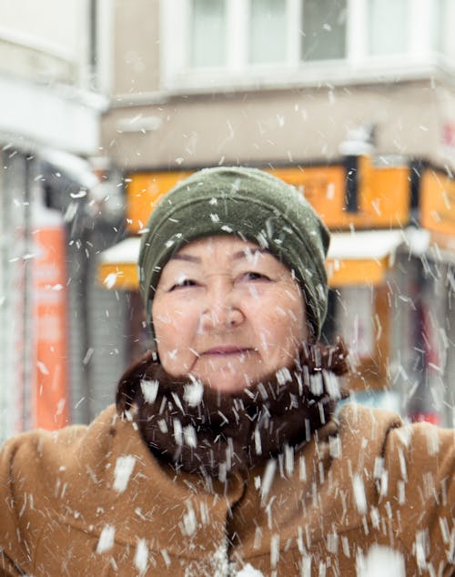 Calm elderly Asian woman in warm outfit standing on street in snowy day and looking at camera