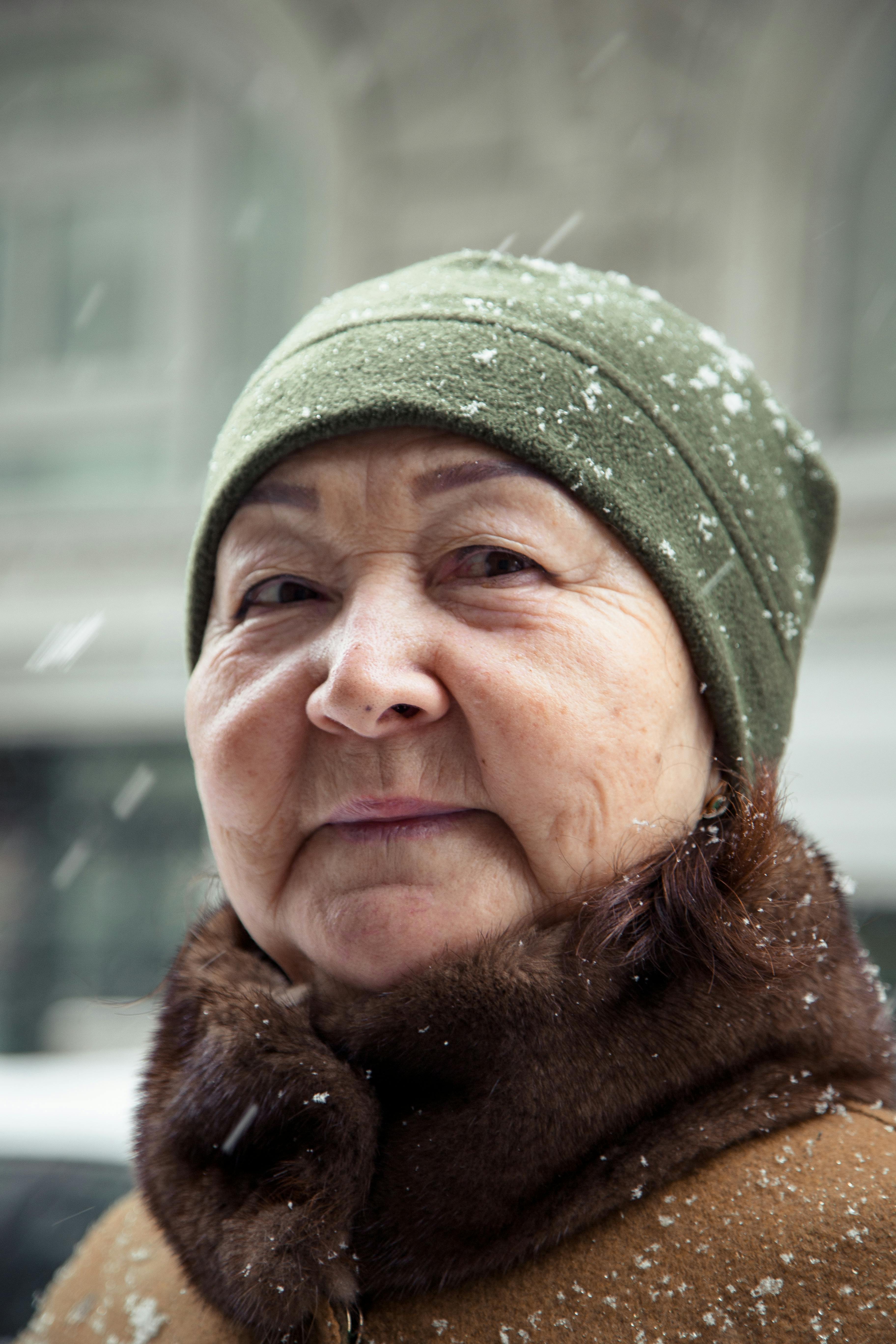 Thoughtful senior Asian woman in warm outerwear looking at camera against blurred buildings on city street in snowy day
