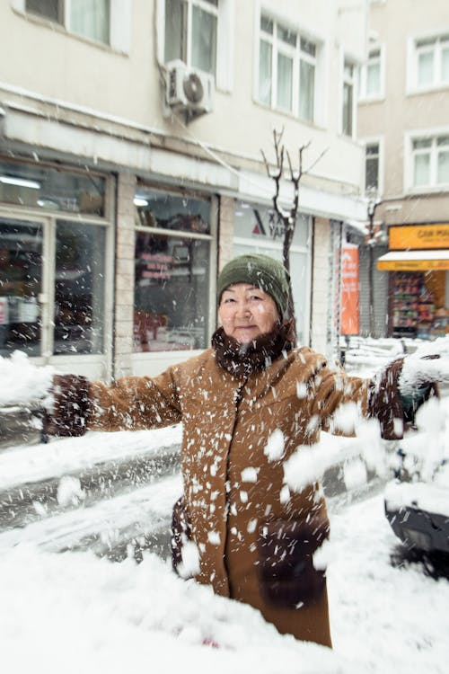 Positive elderly Asian woman in warm outerwear standing and throwing snow on city street in winter time in daylight