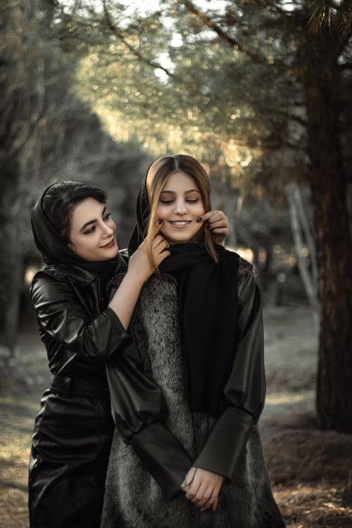 Free Young woman in black outfit with head covered pulling cheeks of mourning girlfriend making smile standing on alley Stock Photo