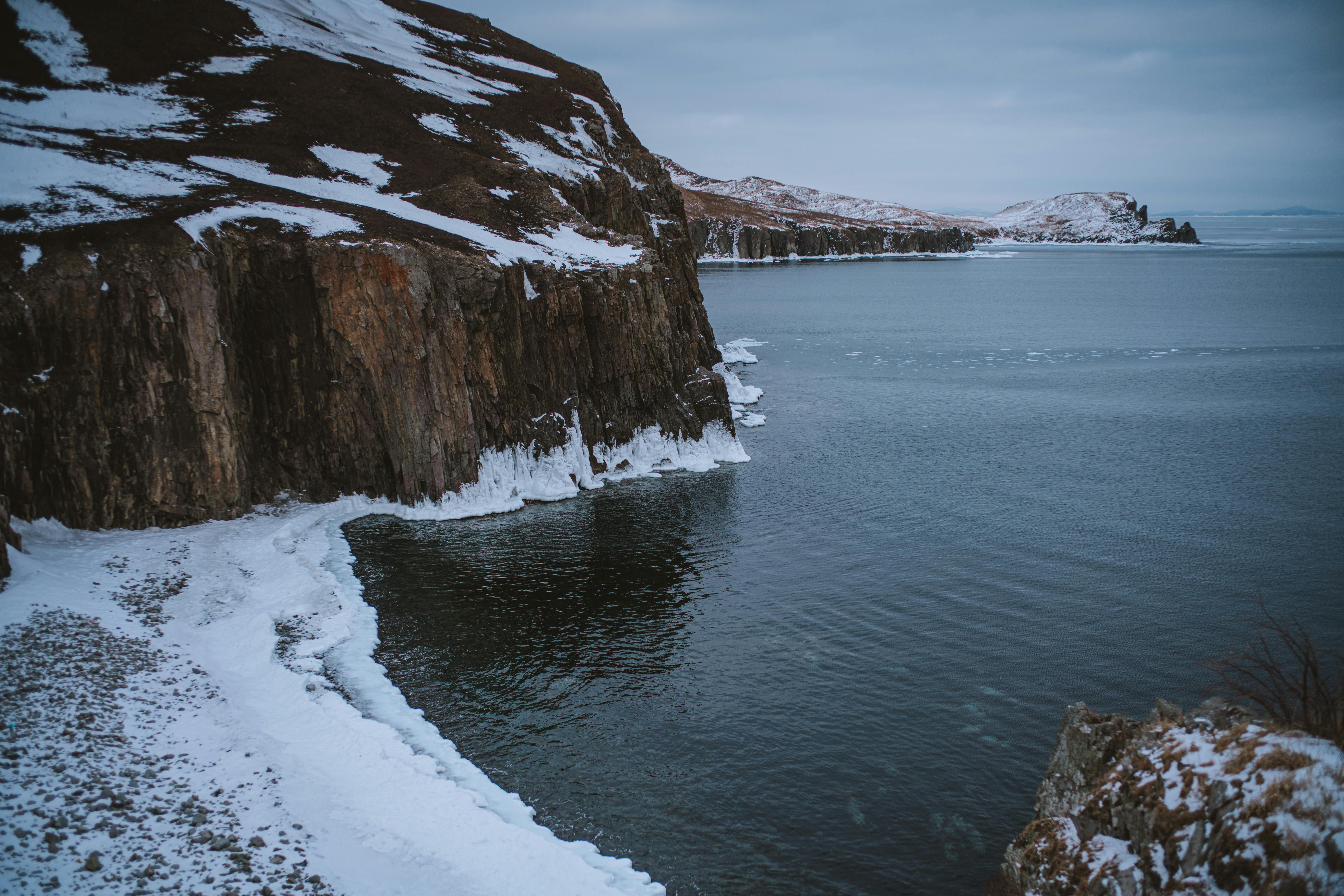 A scenic view of rocky cliffs and snowy shoreline beside a calm winter sea.