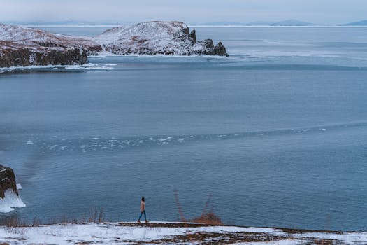 A person walks near a frozen lake surrounded by snowy mountains and calm waters.