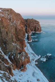 Spectacular aerial view of snow-covered cliffs meeting icy arctic sea in winter twilight.