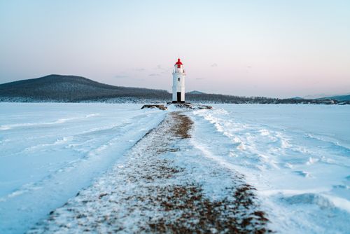 Free A picturesque lighthouse stands amidst a snowy landscape, surrounded by serene mountains at dusk. Stock Photo