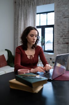 A woman works on her laptop in a cozy home office setting with books and a coffee mug nearby.