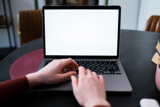 Hands typing on a laptop keyboard with a blank white screen on a round table.