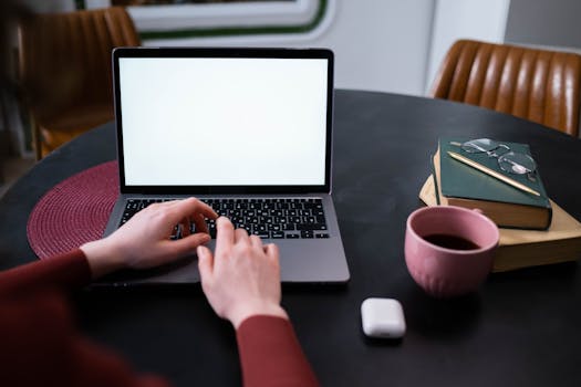 Hands typing on a laptop at a home office setting with coffee, books, and earphones.