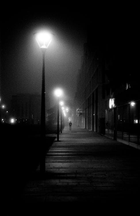Grayscale Photo of a Person Walking on the Sidewalk with Street Lights