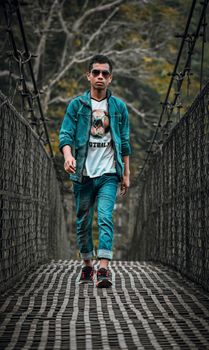 A fashionable young man walks confidently across a suspension bridge in a denim outfit.
