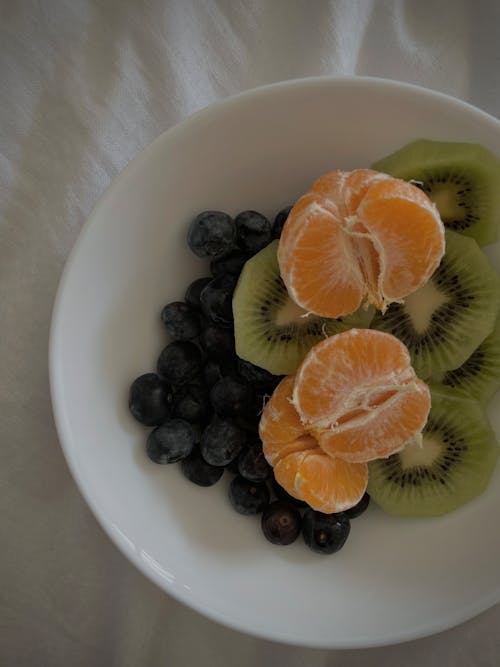 Bowl with mandarins and blueberries with sliced kiwi on bed
