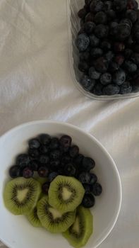 Top view of bowl with ripe tasty blueberries and sliced kiwi near plastic container with berries on white bed