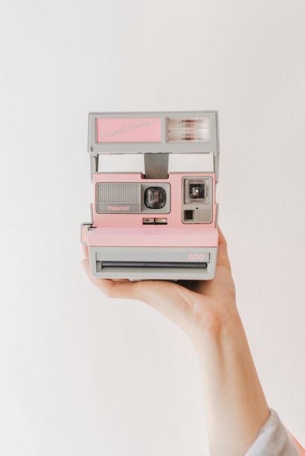 Close-up of a pink vintage Polaroid camera held by a hand on a plain background.